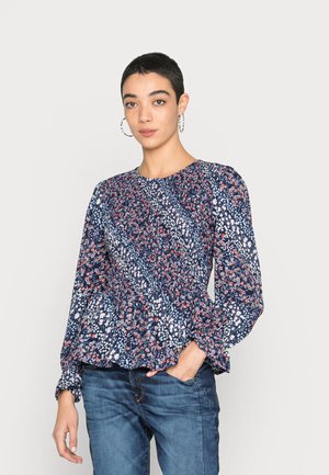 Blusa com padrão floral, mangas longas e bufantes, base azul-marinho e pormenores coloridos em vermelho e branco. A barra peplum adiciona textura. Combinada com jeans de ganga.