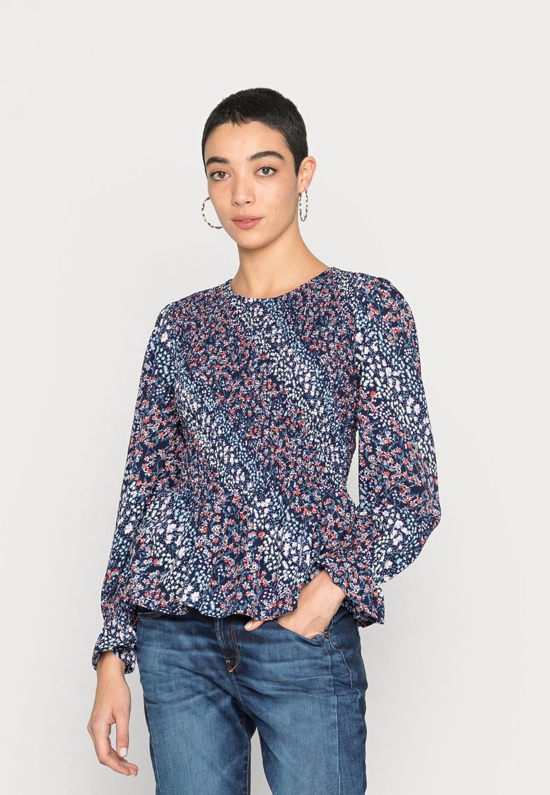 Blusa com padrão floral, mangas longas e bufantes, base azul-marinho e pormenores coloridos em vermelho e branco. A barra peplum adiciona textura. Combinada com jeans de ganga.