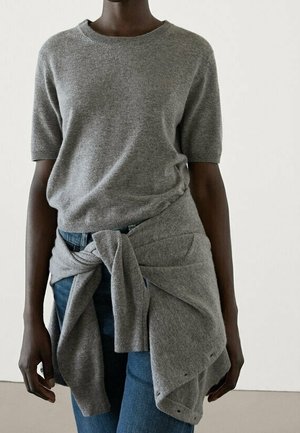 Personne portant un pull à manches courtes gris avec un pull assorti noué autour de la taille, associé à un jean bleu, debout contre un mur uni.