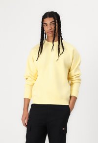 Yellow cotton sweatshirt with a round neckline and ribbed cuffs. Features a small green logo on the chest. Paired with black cargo pants.