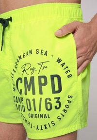 Pantaloni da bagno giallo neon con stampa grafica nera, vita elasticizzata con cordino e tasche laterali. Presenta dettagli testuali audaci.