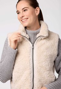 Krēma krāsas mākslīgās kažokādas veste ar augstu apkakli; aprīkota ar priekšējo rāvējslēdzēju un valkāta kopā ar teksturētu pelēku džemperi zem tās. Mīksta, pūkaina tekstūra.