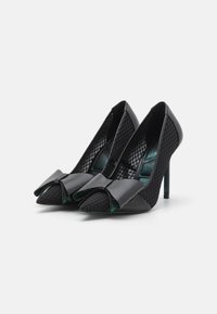 Chaussures à talons hauts noires avec un design en mesh, un bout pointu et un nœud décoratif à l'avant. Elles présentent une finition brillante et une semelle intérieure couleur sarcelle.