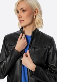 Giubbotto di pelle nera con colletto alto, zip frontale e due tasche frontali con zip orizzontali. Indossato sopra un maglione blu. Texture liscia.