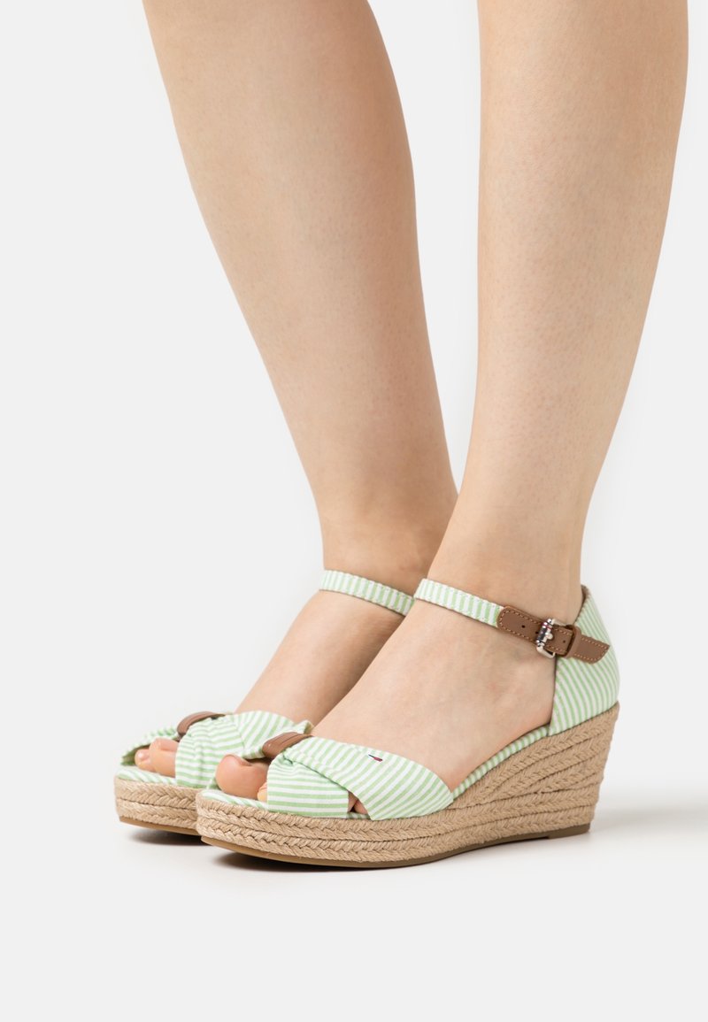 Jambes portant des sandales compensées rayées vert clair et blanc avec brides chevilles et boucles marron sur fond blanc.