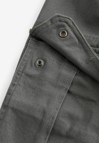Primer plano de una tela gris, que presenta una costura y un detalle de broche de metal. El material es suave con un leve brillo, lo que indica un diseño duradero.