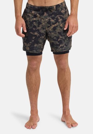 Mężczyzna noszący czarne i brązowe spodenki w kamuflażowy wzór z czarnymi wewnętrznymi szortami kompresyjnymi, stojący boso na białym tle.