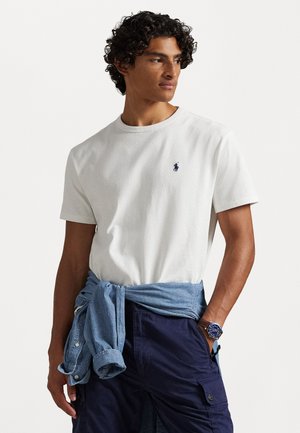 Tricou alb din bumbac cu un logo brodat albastru, asortat cu pantaloni scurți cargo bleumarin și o cămașă din denim albastră legată în jurul taliei.