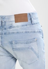 Jean en denim bleu clair avec une poche arrière et un patch de marque en cuir marron portant l'inscription « Opus », porté par une personne en chemise blanche.