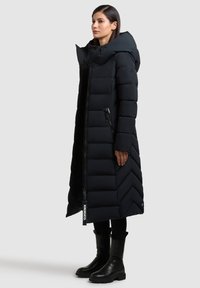 Kvinde iført en lang sort dunjakke med hætte, lynlås foran, diagonal sidesyning og sorte støvler mod en ensfarvet baggrund.