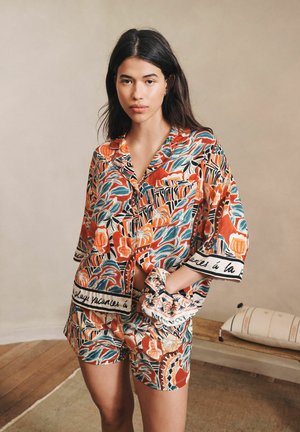 Pikkade tumedate juustega naine, kes kannab värvilist mustrilist lühikeste varrukatega särki ja sobivaid lühikesi pükse, hoides peapaela, seistes siseruumis puitpõrandal.