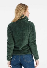 Grön fleece-pullover med hög krage, tillverkad av ett mjukt, texturerat material. Baksidan visar en figursydd design och ribbade muddar.