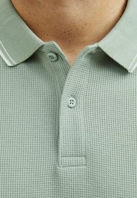 Lys grøn polo skjorte lavet af tekstureret stof med kravehals, to knapper og hvide stribede detaljer på kraven.