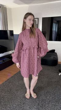 Rosa klänning med mönster, volang vid midjan, ballongärmar och hög krage. Bärs med beige sandaler på en texturerad matta.