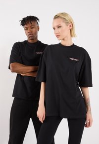 Black oversized t-shirts made of cotton, featuring a small logo on the chest. Modelled on two individuals with relaxed fits.