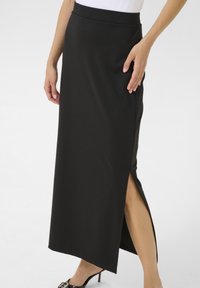 Jupe maxi noire, ajustée, avec une fente haute sur un côté. Fabriquée en tissu lisse avec une taille classique, associée à des chaussures à talons hauts.