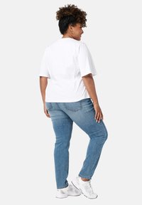 Witte katoenen T-shirt met korte mouwen, gecombineerd met lichtblauwe jeans en witte sneakers. Achtergrond is neutraal grijs.