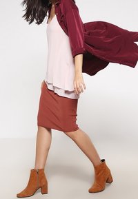 Vrouw wandelt in een gelaagde roze top, een roestkleurige pencil skirt, roestkleurige suède enkellaarsjes en een vloeiende donkerrode jas.