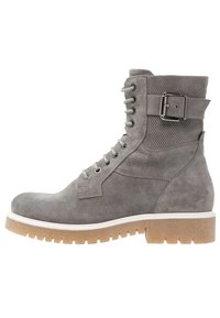 Harmaa mokkanahkasaapas nilkkapituudella, jossa on paksu kumipohja, nauhakiristys edessä ja sivussa solk yksityiskohta. Teksturoitu päällinen, jossa on sileitä aksentteja.