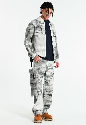 Joven con rastas que lleva chaqueta y pantalones grises con un patrón abstracto, camisa azul marino, zapatos marrones y una larga correa con estampado que cuelga de la cintura.