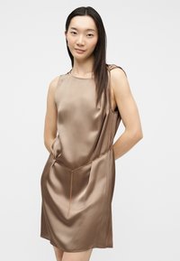 Γυναίκα με μακριά σκούρα μαλλιά φοράει αμάνικο, λαμπερό σατέν φόρεμα σε γκριζωπό τόνο με λεπτή χρυσή αλυσίδα στη μέση, σε απλό φωτεινό φόντο.