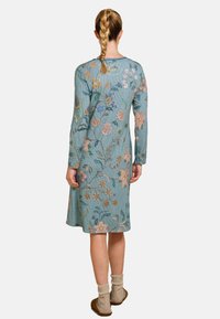 Woman with braided hair wearing a long-sleeve, knee-length teal dress with multicolored floral patterns, beige socks, and brown slippers, standing.