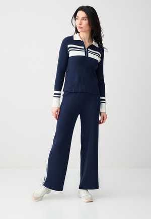 Donna in piedi che indossa pantaloni a coste blu navy e maglione abbinato con zip e strisce bianche, abbinati a scarpe da ginnastica bianche, guarda di lato.