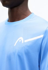 Κοντινό πλάνο ενός ατόμου που φοράει ανοιχτό μπλε αθλητική φανέλα με ένα λευκό αφηρημένο λογότυπο στο στήθος και αθλητική υφή.