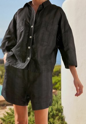 Lockere schwarze Button-Down-Bluse mit Vorderentaschen, kombiniert mit passenden schwarzen Shorts, getragen von einer stehenden Person im Freien in der Nähe von Grünflächen.
