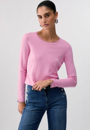 Donna con capelli scuri raccolti in uno chignon, che indossa orecchini a cerchio d'argento, un top rosa a maniche lunghe e jeans blu a vita alta con bottoni laterali.