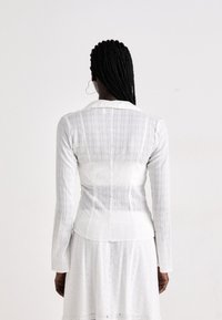 Black woman with long braided hair wearing white textured long-sleeve shirt and skirt, standing facing a plain light background.