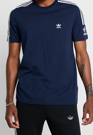 Homme portant un t-shirt Adidas bleu marine avec des bandes blanches sur les épaules et le logo sur la poitrine et la manche, assorti à un pantalon Adidas noir avec logo.