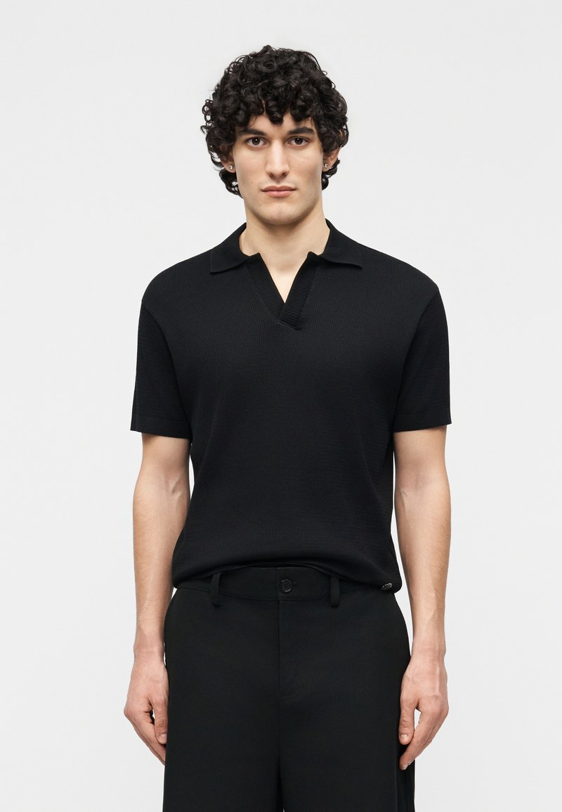 Jeune homme aux cheveux bouclés portant une chemise à col en maille noire à manches courtes et un pantalon noir, debout devant un fond clair uni.