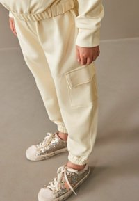 Cremefarvede joggingbukser med elastiske manchetter og en sidelomme, parret med sneakers dækket af glimmer og hvide snørebånd.