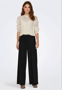 Pullover en maille côtelée beige clair avec un col rond et des manches retroussées, associé à un pantalon noir à jambe large avec une taille élastique.