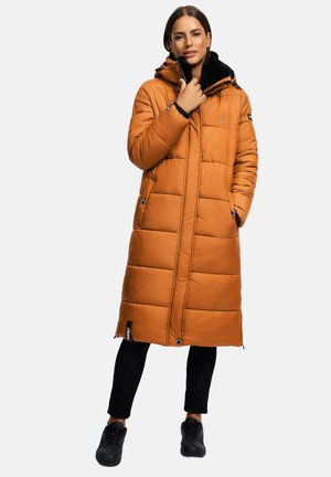 Woman wearing a long, orange puffer coat with black fur lining, black pants, and black sneakers, standing with one hand in pocket.
