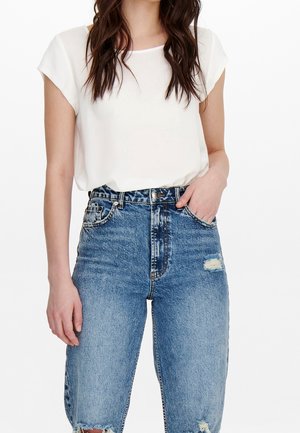 Femme portant un chemisier blanc à manches courtes rentré dans un jean bleu déchiré taille haute, avec une main dans la poche, sur fond blanc.