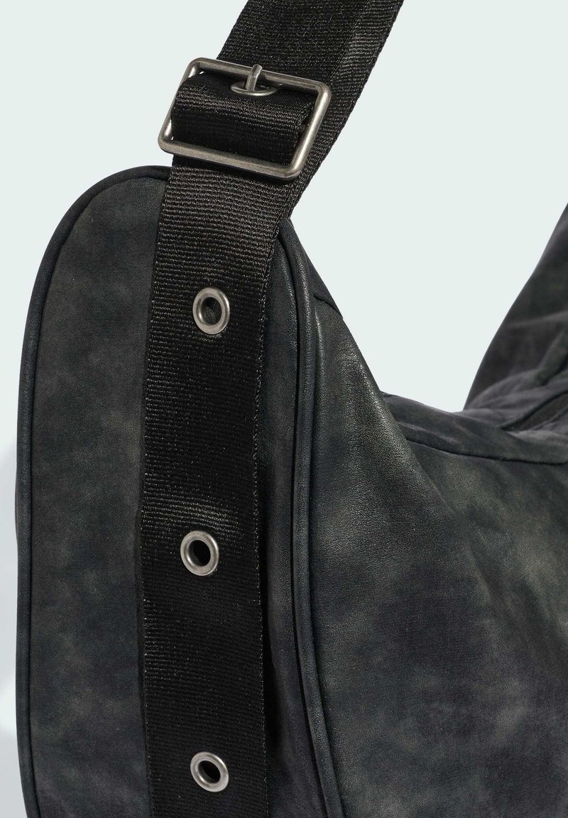 Schwarze Ledertasche mit verstellbarem schwarzen Stoffriemen, Metall Schnalle und Ösen, vor einfachem hellem Hintergrund gezeigt.