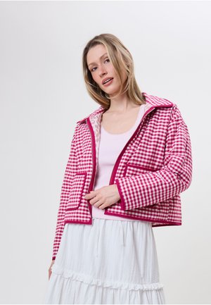 Ung kvinde iført en pink og hvid ternet jakke over en lyserød top og hvid nederdel, stående mod en ensartet lys baggrund.