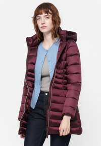 Bordeaux pufferjack met rits en capuchon, met een geborduurd patroon, gedragen over een lichtblauwe cardigan en een grijze top.