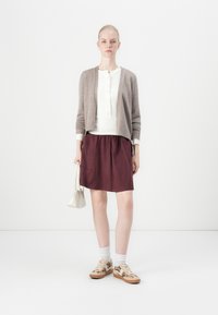 Gebreide bruine cardigan over een witte blouse, gecombineerd met een bordeauxrode rok, witte sokken en sneakers met een patroon, en een kleine handtas in de hand.