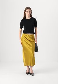 Svart strikket topp med kort erme, gul sateng maxi-skjørt, svarte lakksko med høye hæler, og en liten svart håndveske. Jevn tekstur og tettsittende design.