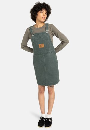 Green denim overall dress with adjustable straps, front pocket, and chest patch. Worn over a long-sleeve gray shirt, paired with black shoes.