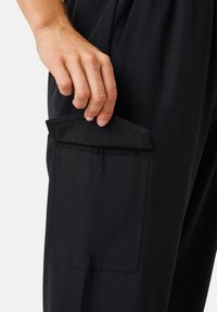 Black trousers with a large patch pocket. The pocket has a fold-over flap and vertical stitching along the edges. Smooth fabric texture.