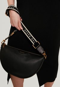 Sac à main en cuir noir à forme demi-lune, doté d'une fermeture éclair dorée et d'une bandoulière avec l'inscription "TED BAKER" en blanc. Texture lisse et design minimaliste.