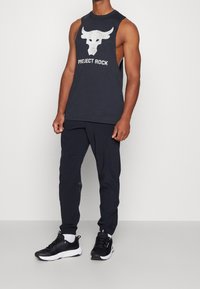 Musta hihaton hihaton t-paita, jossa on härkälogo ja "PROJECT ROCK" -teksti, yhdistettynä mustiin urheiluhousuihin ja mustiin kenkiin, joissa on valkoisia yksityiskohtia.