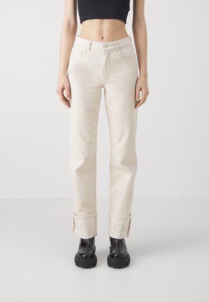 Jeans beige a vita alta con gambe dritte, risvoltati alla caviglia. Il tessuto appare leggermente testurizzato. Indossati con stivaletti neri con plateau.