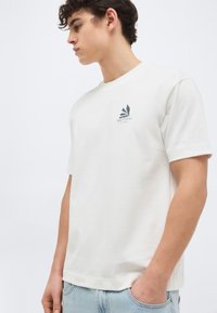 Νεαρός άνδρας με λευκό πουκάμισο κοντομάνικο με μικρό λογότυπο στο στήθος, φοράει ανοιχτόχρωμο τζιν και στέκεται με το ένα χέρι στην τσέπη.