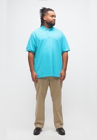 Άνδρας με dreadlocks που φοράει τουρκουάζ πόλο μπλούζα, μπεζ παντελόνια και μαύρα παπούτσια, στέκεται και κοιτάζει στο πλάι σε απλό λευκό φόντο.