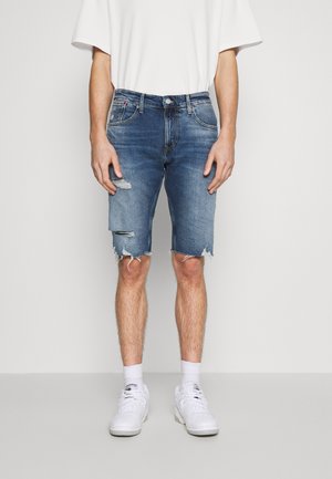 Denim-Shorts in verblasstem Blau mit Used-Effekten, fransigem Saum und Fünf-Taschen-Design, kombiniert mit einem weißen T-Shirt und weißen Sneakern.